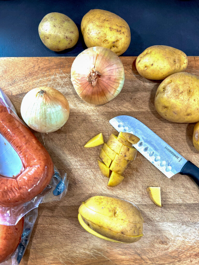 Sausage, onion, knife, cubed and whole potatoes on a cutting board.